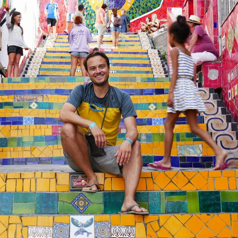 Lachender Mann sitzt in kurzer sommerlicher Kleidung auf ein farbigen Treppe aus vielen farbigen Steinen