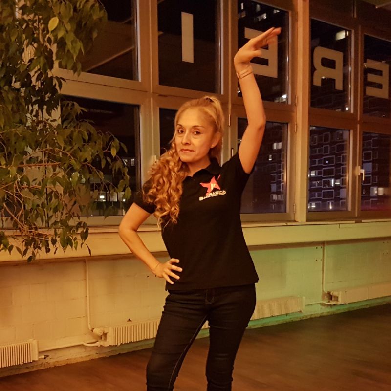 Frau in schwarzer Kleidung steht mit hohen SChuhen auf der Tanzfläche und streckt den Arm in die Höhe zu einer Tänzerischen Pose.