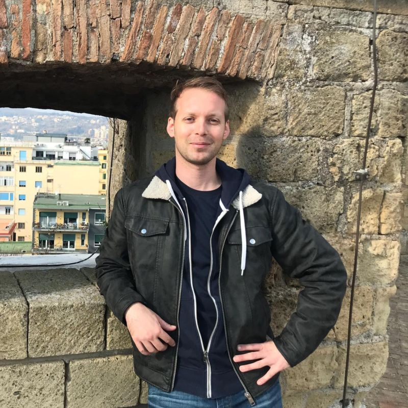 Mann steht vor einer Steinmauer auf einer Burg und hat eine schwarze Jacke an