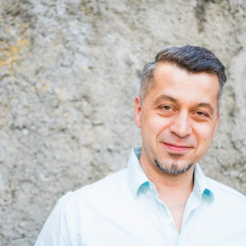 Mann mit blauem Hemd und kurzen Harren steht lächelnd vo einer Steinwand