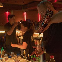 Ein Mann beim Cocktail mixen und eine Frau beim kassieren, sowie ein anderer Mann der aufräumt