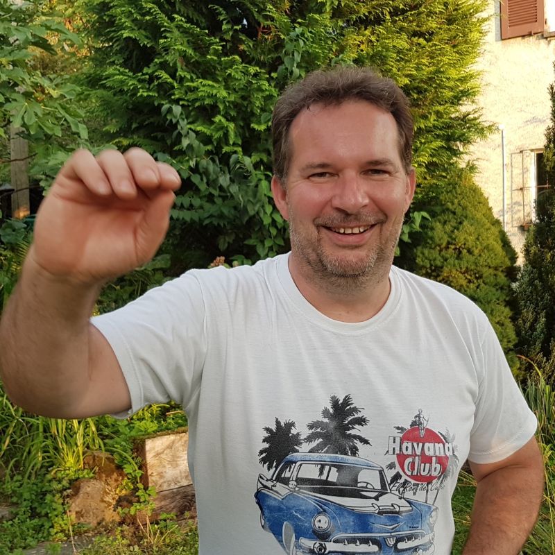 Mann mit kurzen Haaren mit weissem T-Shirt mit Havana club Aufschrift im Garten, welcher mit der Hand winkt