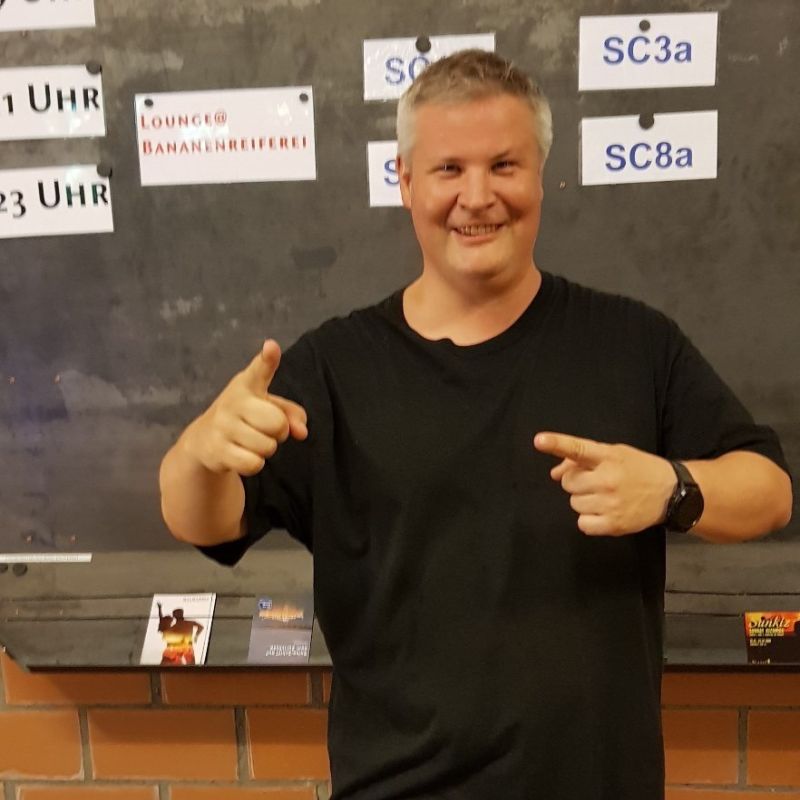 Mann in schwarzem Shirt steht vor einer Wand und zeigt mit den Fingern. Er lacht und hat gute Laune