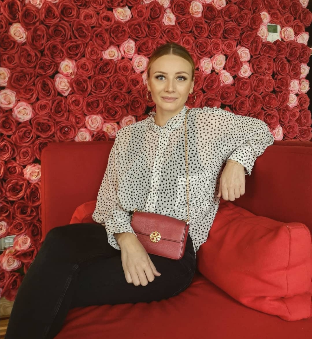 Junge Frau auf einem roten Sofa mit schwarzer Hose und weisser Bluse mit Rosenbild im Hintergrund