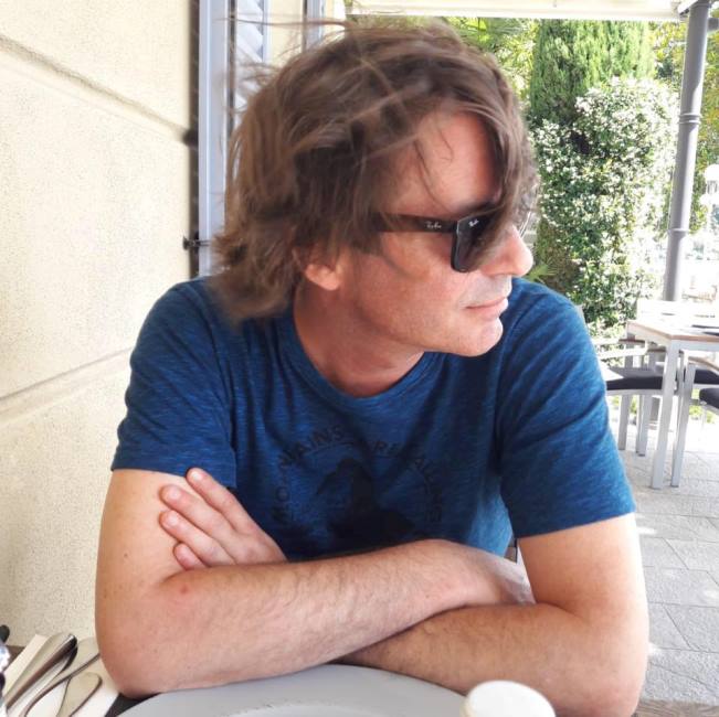 Mann mit dunkler Sonnenbrille und halblangen braunen Haaren an einem Tisch sitzend mit verschränkten Armen von der Seite