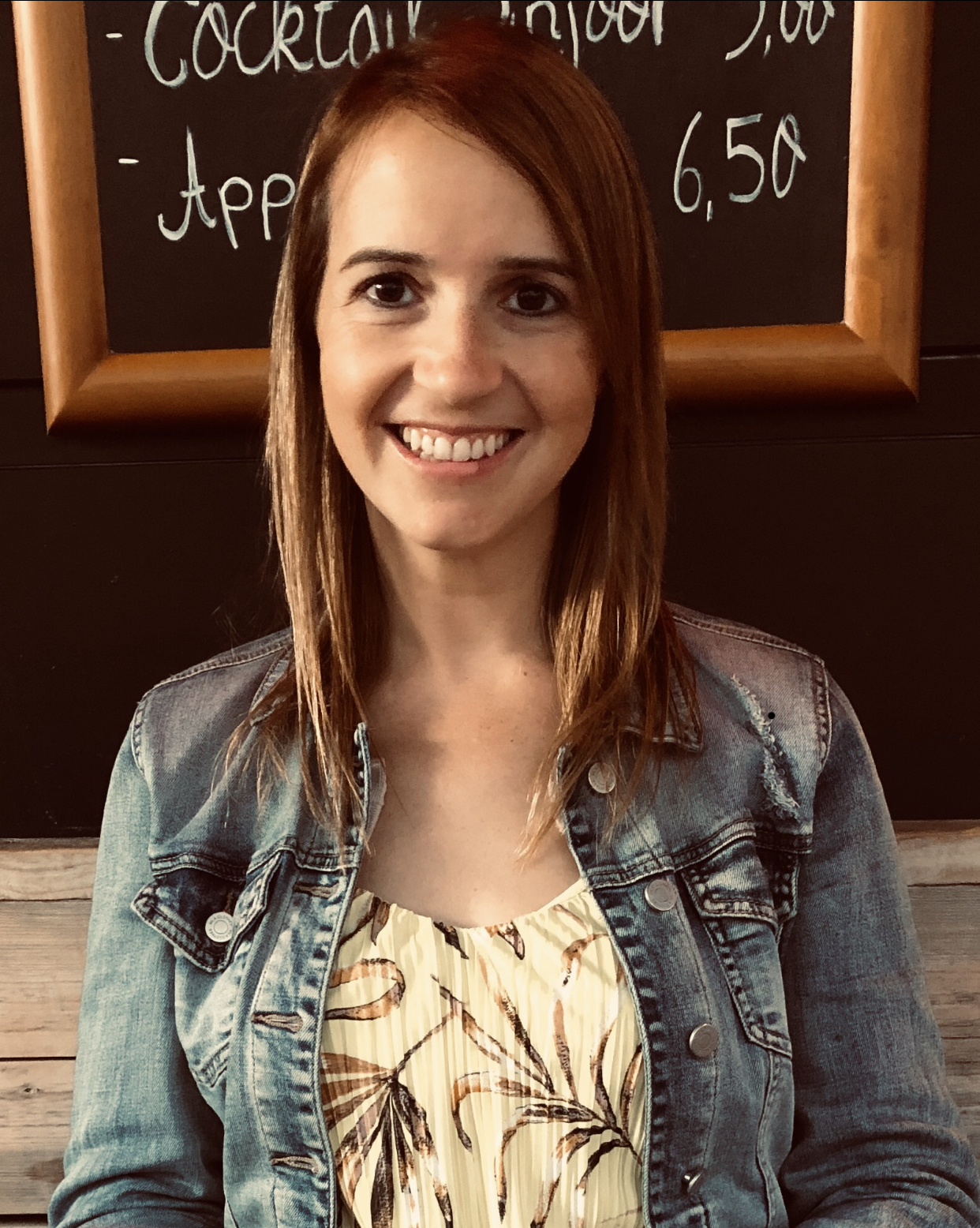 Lachende Frau mit offenen langen Haaren sitzt auf einer Bank und trägt eine Jeansjacke mit heller Bluse