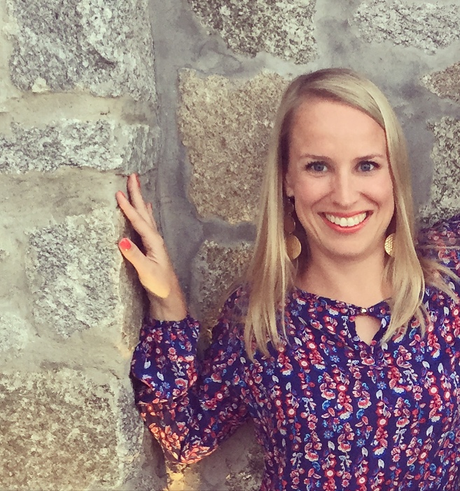 Frau mit langen blonden Haaren steht vor einer Steinwand und lächelt in die Kamera. Eine Hand berührt die Wand