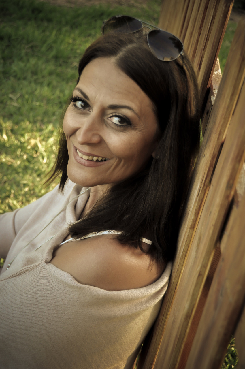 Frau mit braunen langen Haaren trägt beiges Oberteil und eine Sonnenbrille im Haar. Sie lehnt sich an einen Holzzaun an