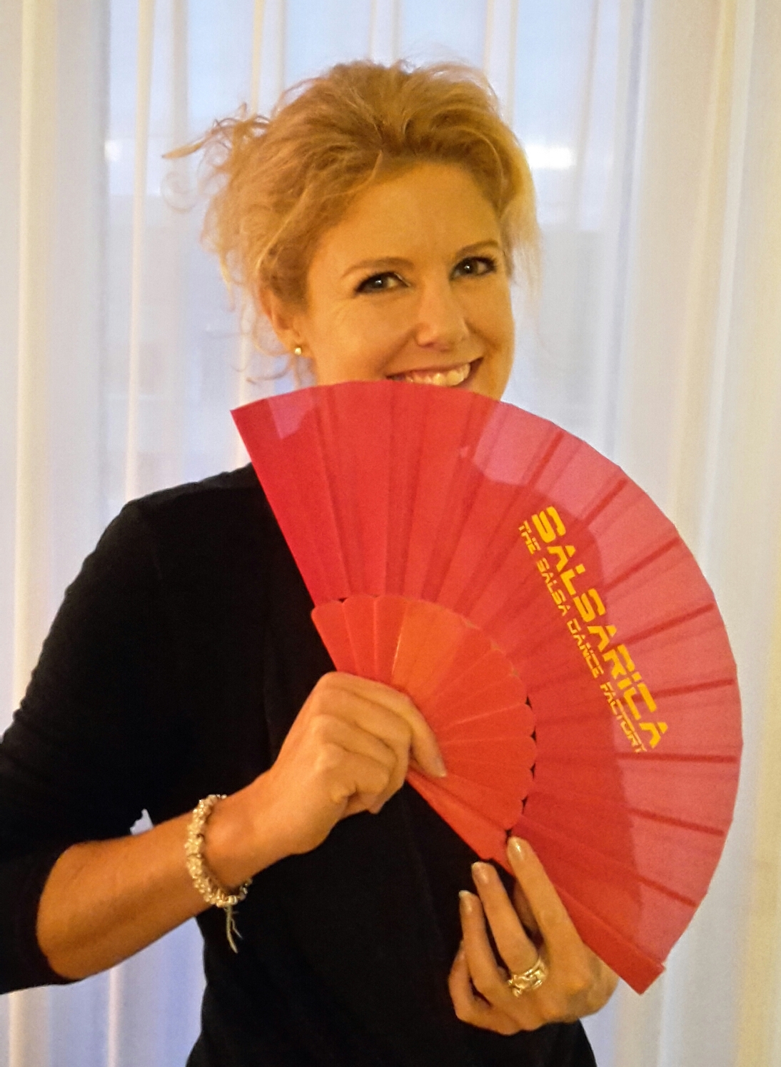 Frau mit blondem Haar und schwarzen Oberteil schaut hinter einem roten Fächer von SalsaRica hervor und lächelt