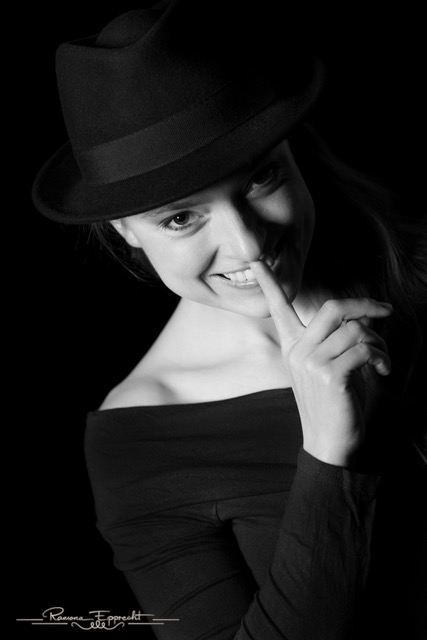 Frau auf einer schwarz/weiss Aufnahme schaut in die Kamera und trägt einen schwarzen Hut. Sie lächelt und deutet mit dem Finger an leise zu sein