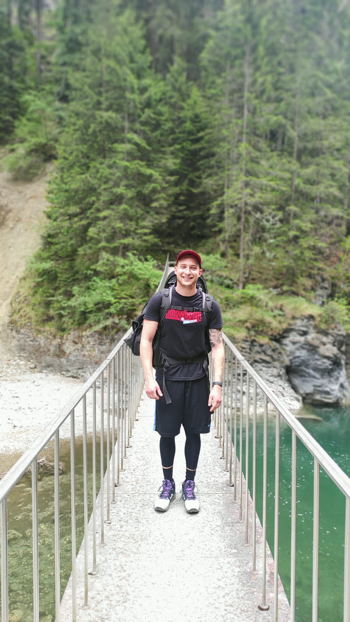Mann steht auf einer Brücke in Wanderkleidung und einem Hut auf dem Kopf, er lacht und ist fröhlich