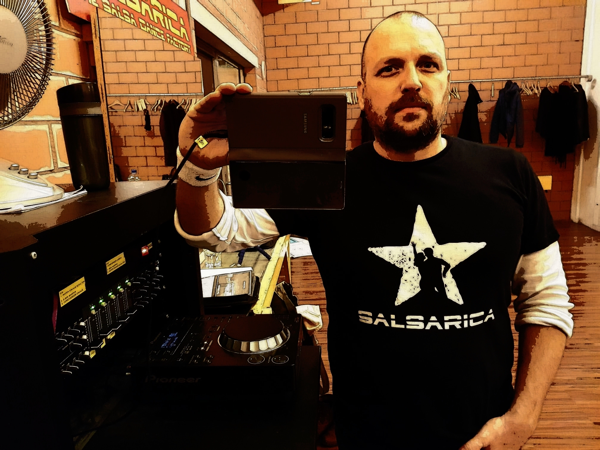 Mann im schwarzen Salsarica Shirt steht bei der Musikanlage und macht von sich selbst mit dem Handy ein Foto