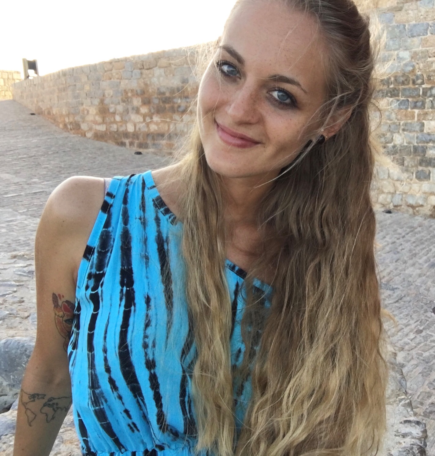 Frau mit langen blonden offenen lockigen Haaren steht am Hafen und trägt ein blaues Kleid. Sie lächelt und ihre blauen Augen leuchten