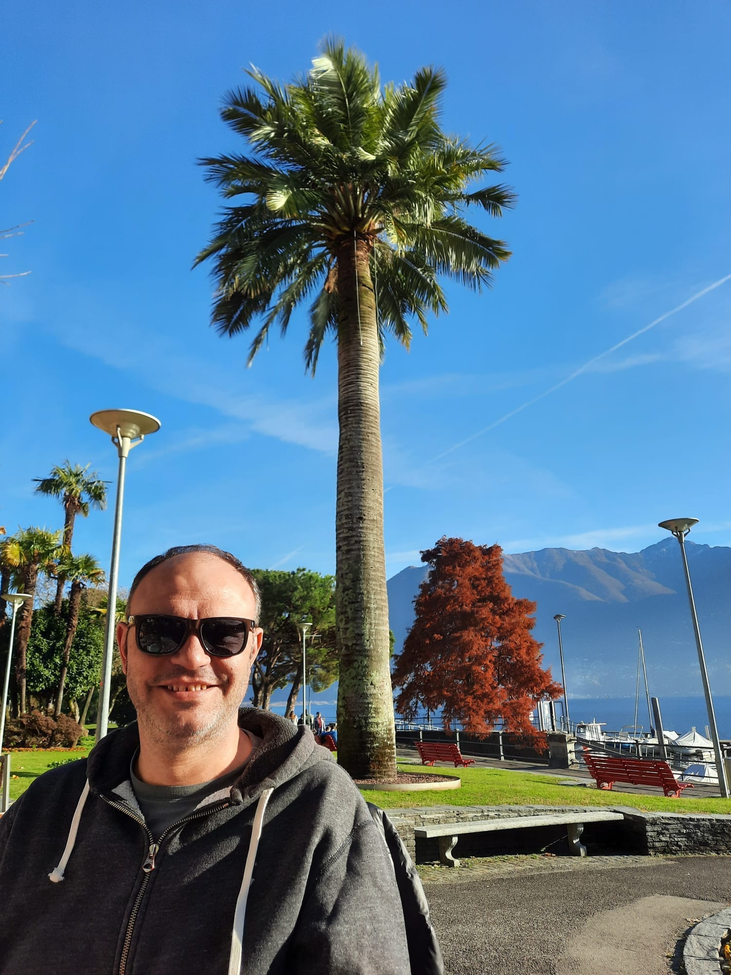 Mann steht mit Sonnenbrille und Sweatjacke im Tessin in der Sonne und lächelt
