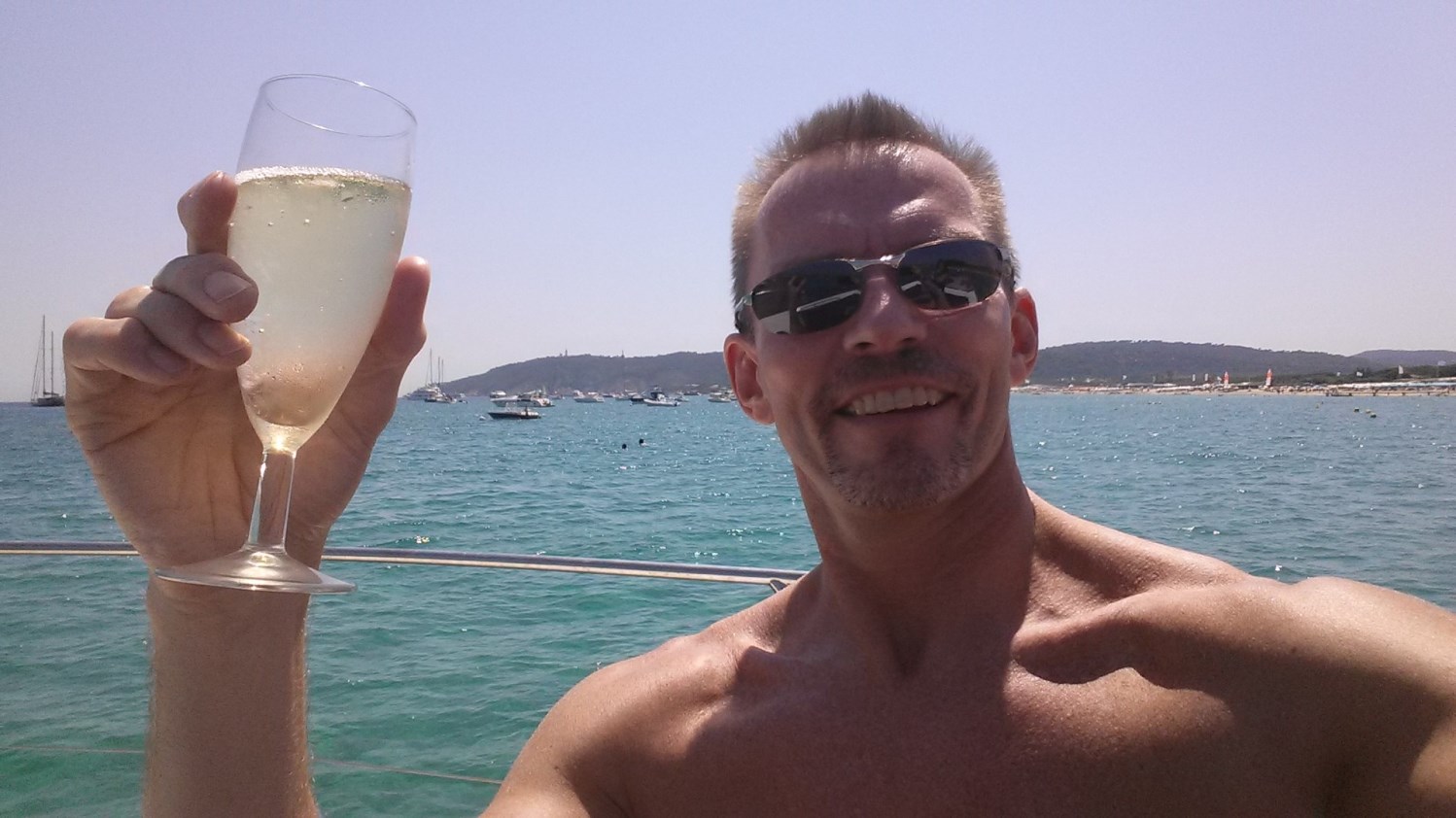 Mann mit nackten Oberkörper steht auf einem Schiff und hält ein Glas Sekt in die Kamera.