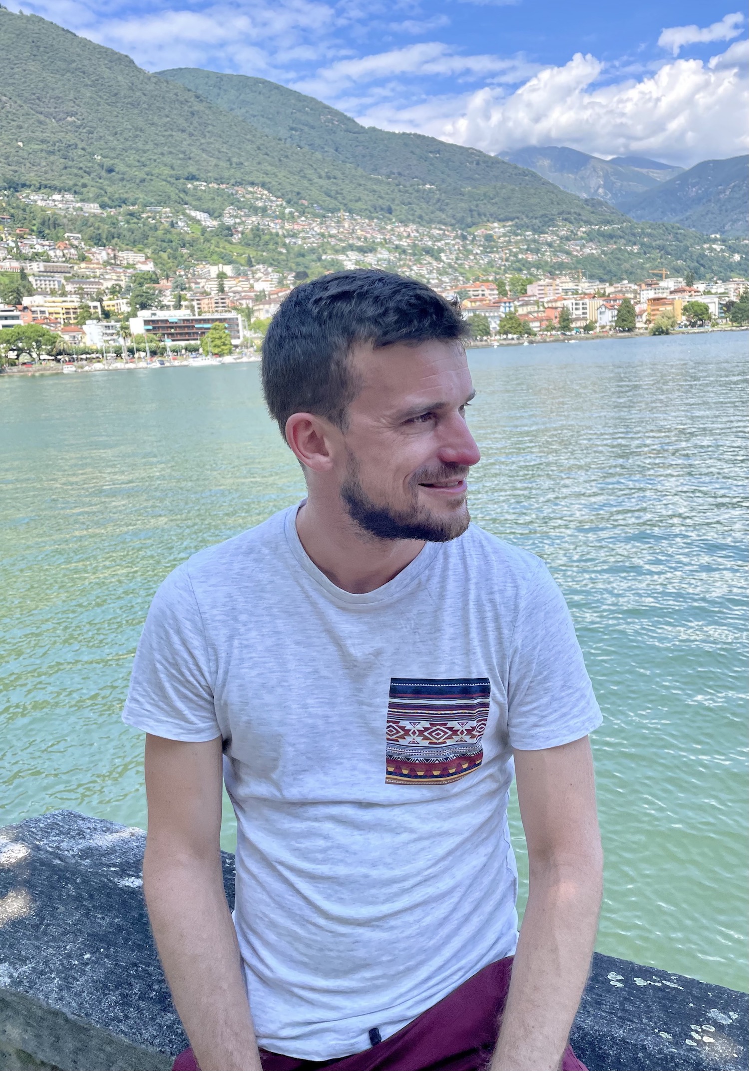 Mann sitzt am Ufer und schaut auf die Seite. Er trägt ein Shirt und lächelt