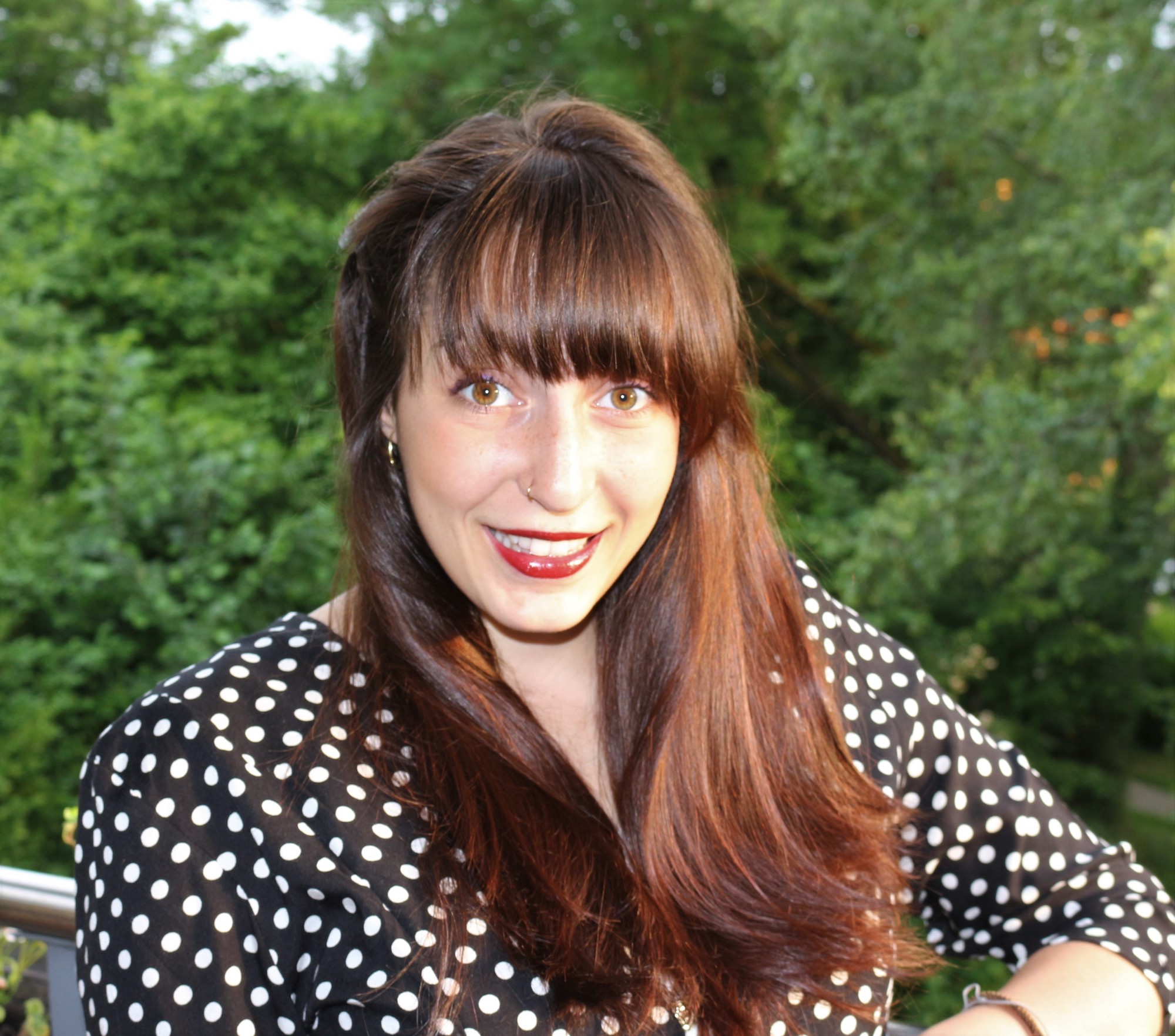 Frau mit rötlichem langen offenen Haaren und einer weiss gepunkteten Bluse steht vor grünen Bäumen