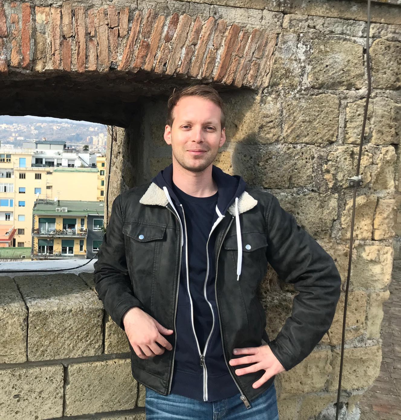 Mann steht vor einer Steinmauer auf einer Burg und hat eine schwarze Jacke an